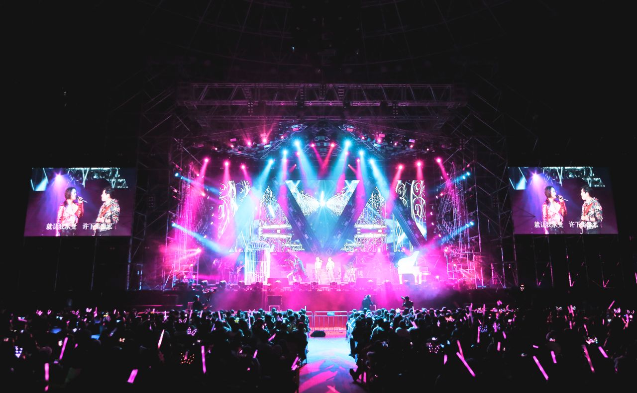Anonymous artists with microphones giving performance on platform illuminated by colorful spotlights in concert hall with audience at night